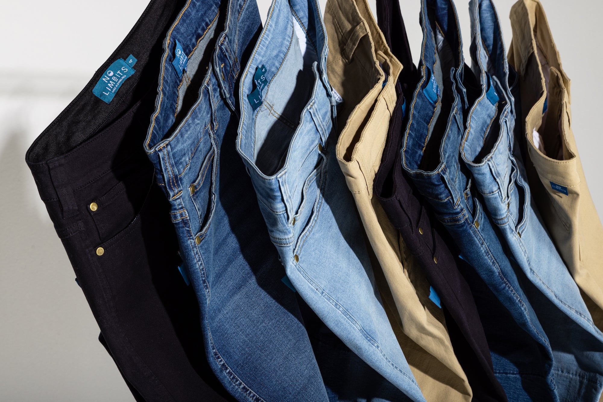 Jeans of multiple different colors are hung together overlapping one another. The colors are black, dark blue, light blue, and khaki. 