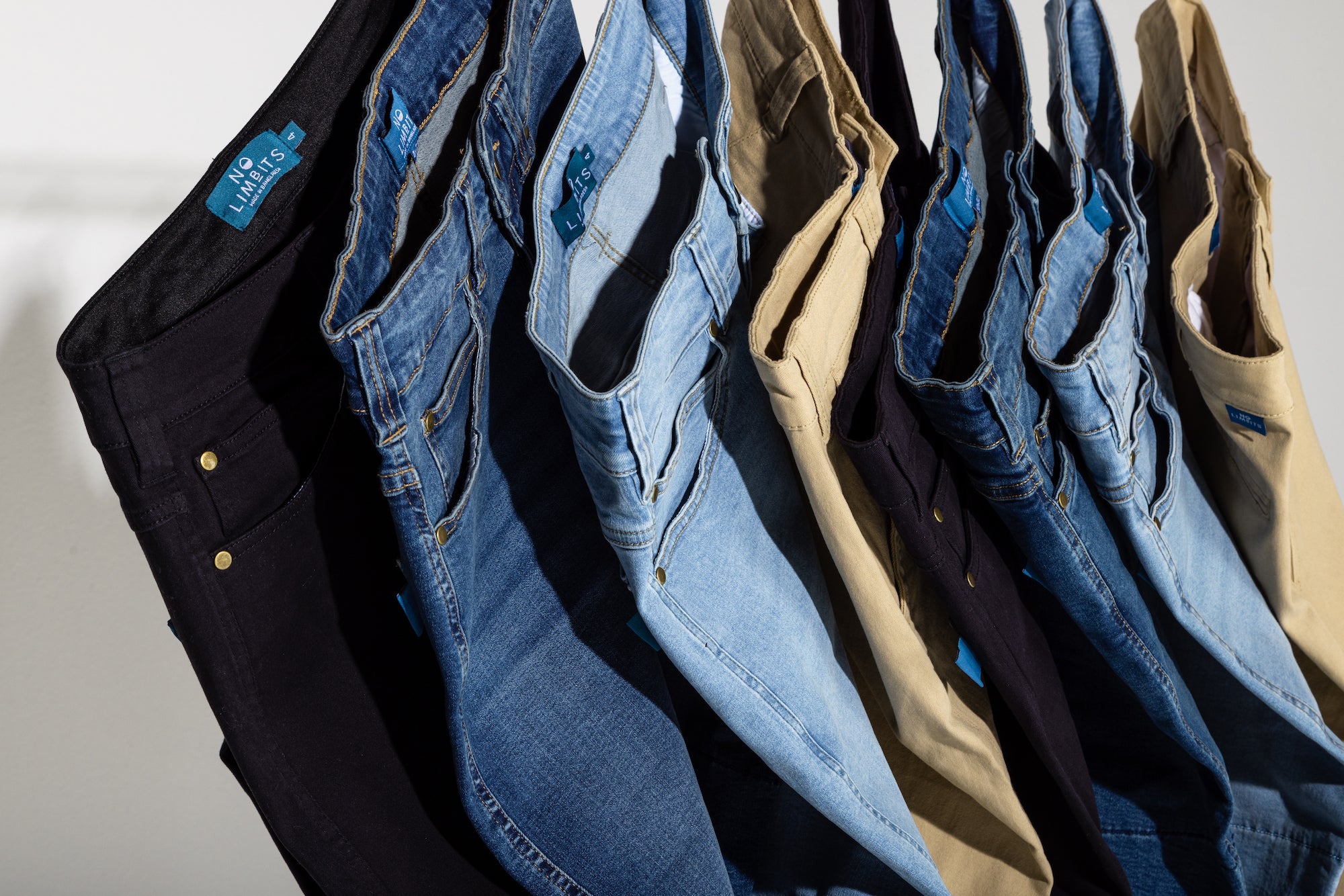 Jeans of multiple different colors are hung together overlapping one another. The colors are black, dark blue, light blue, and khaki.