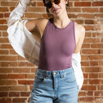 A white woman with black ponytail, brown sunglasses, and hoop earrings wears a mauve tank top and denim jeans. A white jacket is off her shoulders. She's by a brick background.