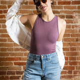 A white woman with black ponytail, brown sunglasses, and hoop earrings wears a mauve tank top and denim jeans. A white jacket is off her shoulders. She's by a brick background.