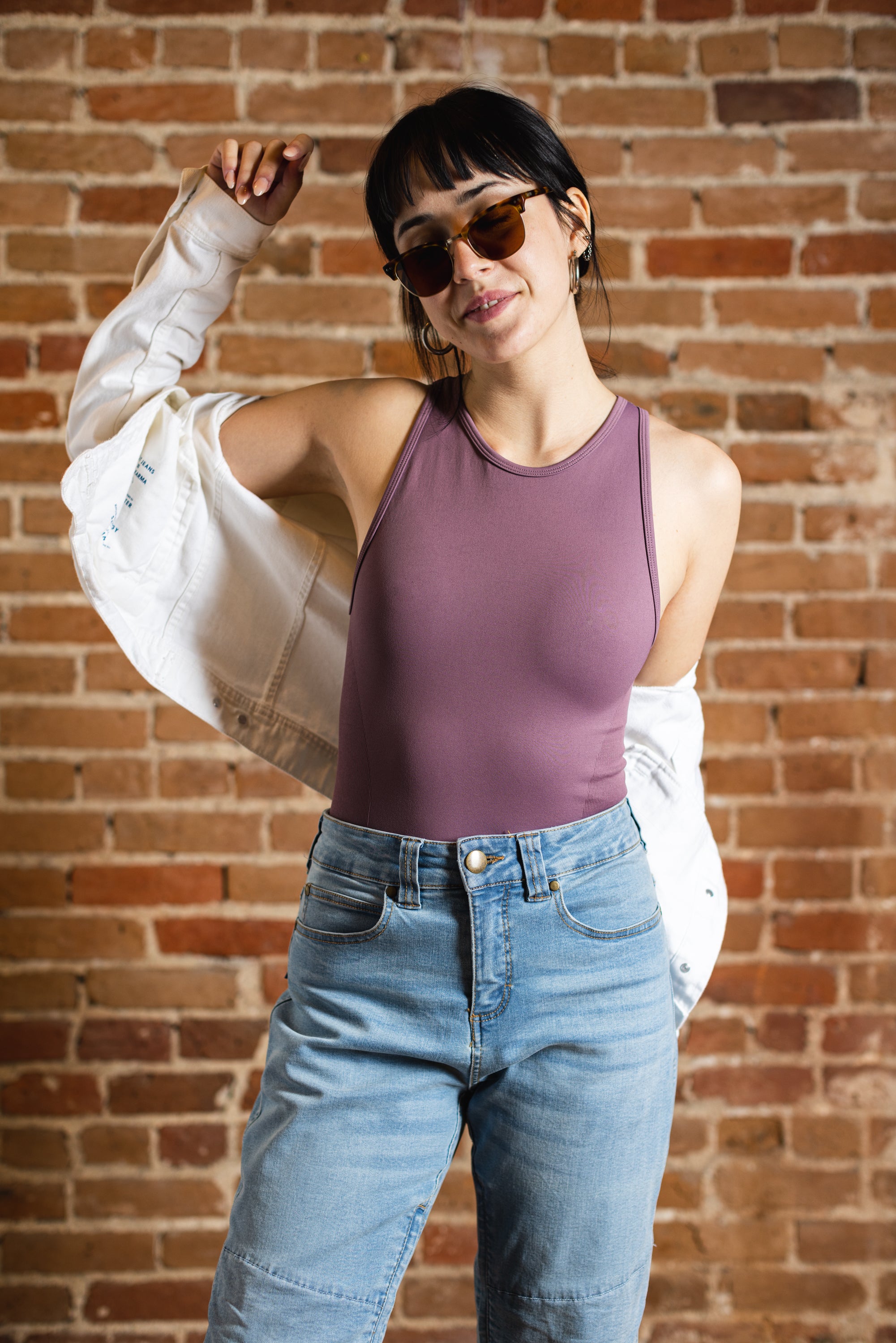 A white woman with black ponytail, brown sunglasses, and hoop earrings wears a mauve tank top and denim jeans. A white jacket is off her shoulders. She's by a brick background.