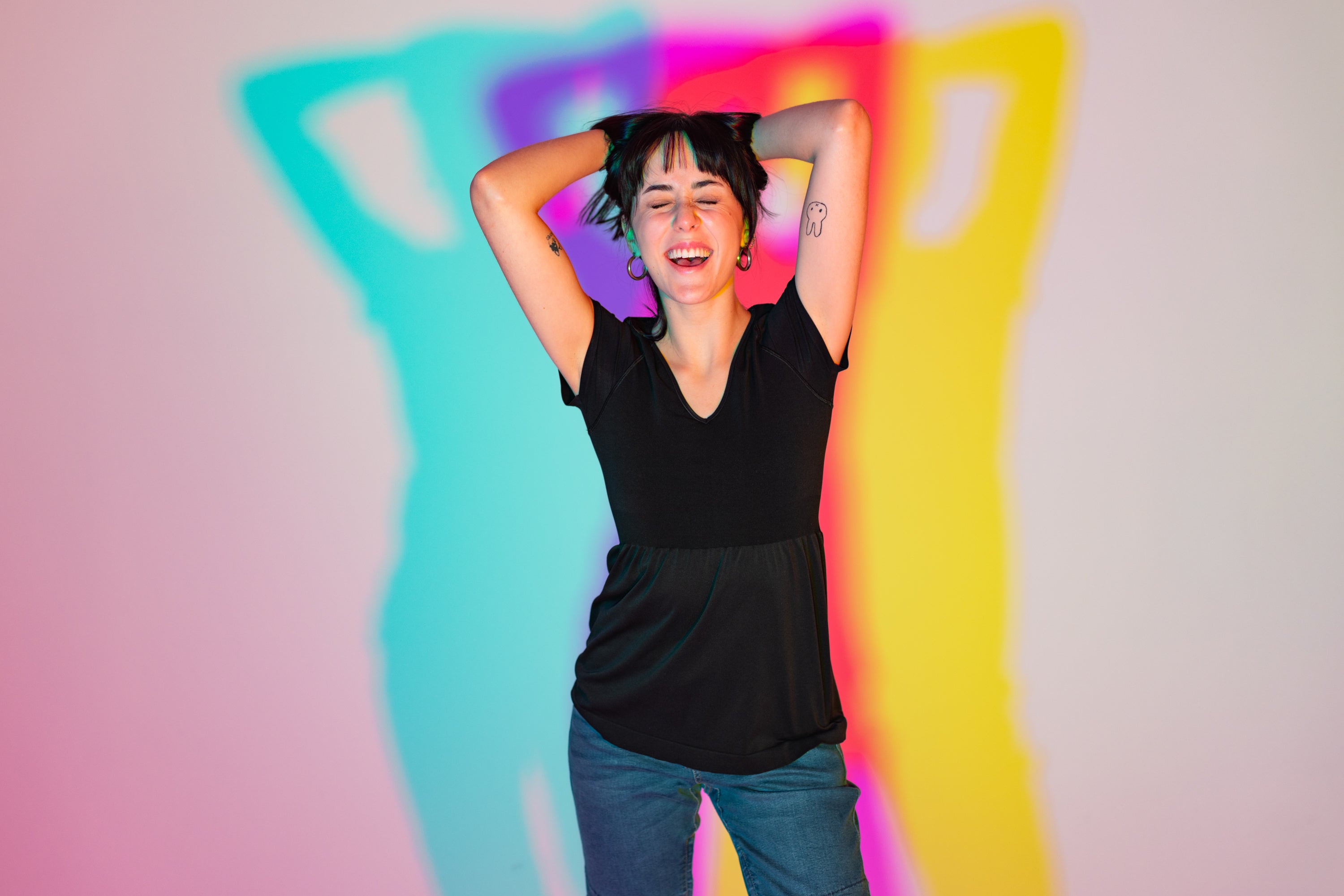 A photo of a woman with dark black hair. She is posing and playing with her hair. She is wearing a black blouse. There is blue and yellow lights flashed behind her.