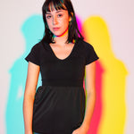Sophia (A woman with long black hair) poses in a white wall. There are outlines behind her that are light blue, purple, pink, and yellow. She is wearing the No Limbits Adaptive Women's Black Sensory Blouse with denim jeans.