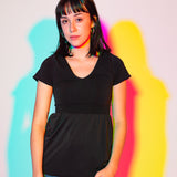 Sophia (A woman with long black hair) poses in a white wall. There are outlines behind her that are light blue, purple, pink, and yellow. She is wearing the No Limbits Adaptive Women's Black Sensory Blouse with denim jeans.