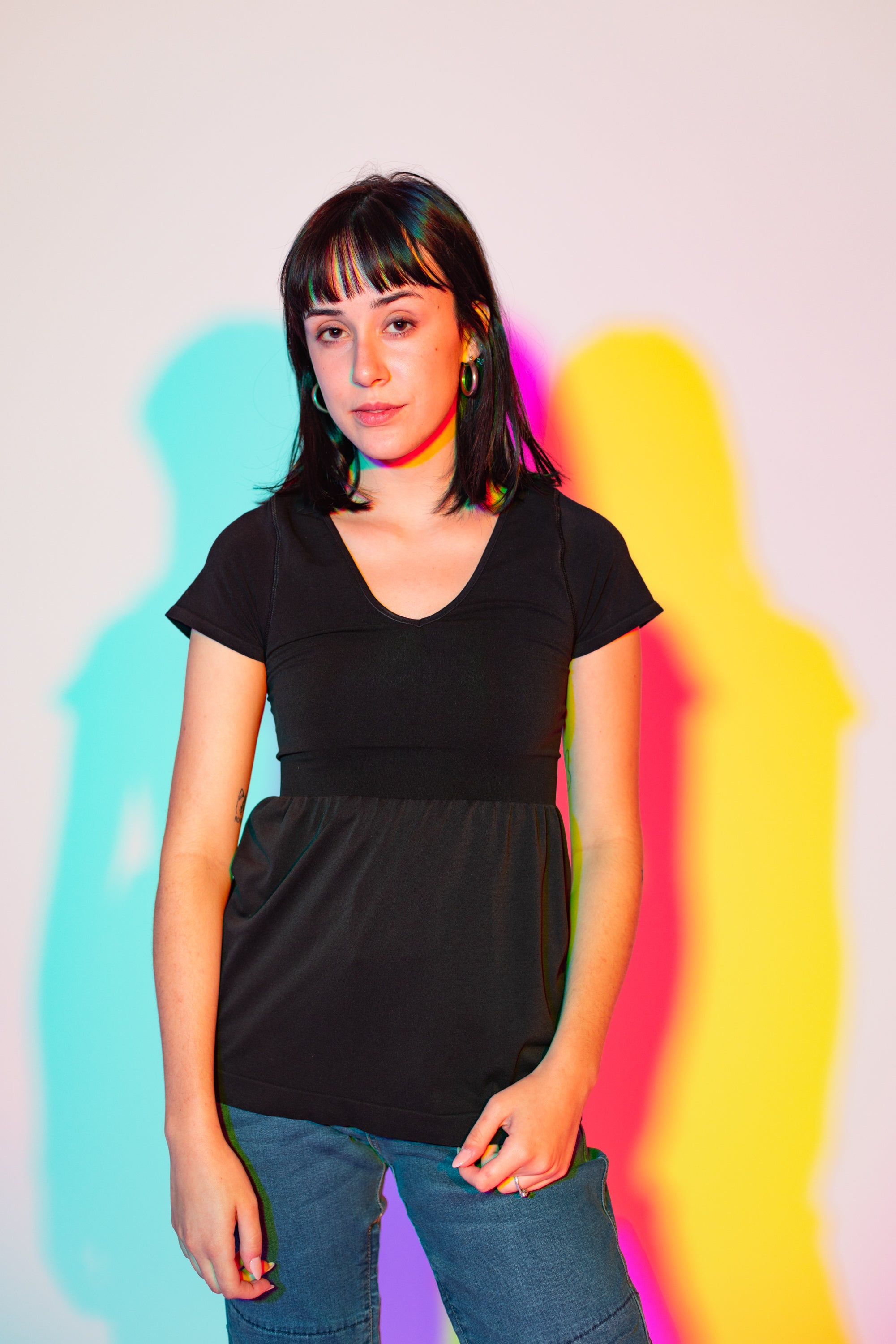 Sophia (A woman with long black hair) poses in a white wall. There are outlines behind her that are light blue, purple, pink, and yellow. She is wearing the No Limbits Adaptive Women's Black Sensory Blouse with denim jeans.