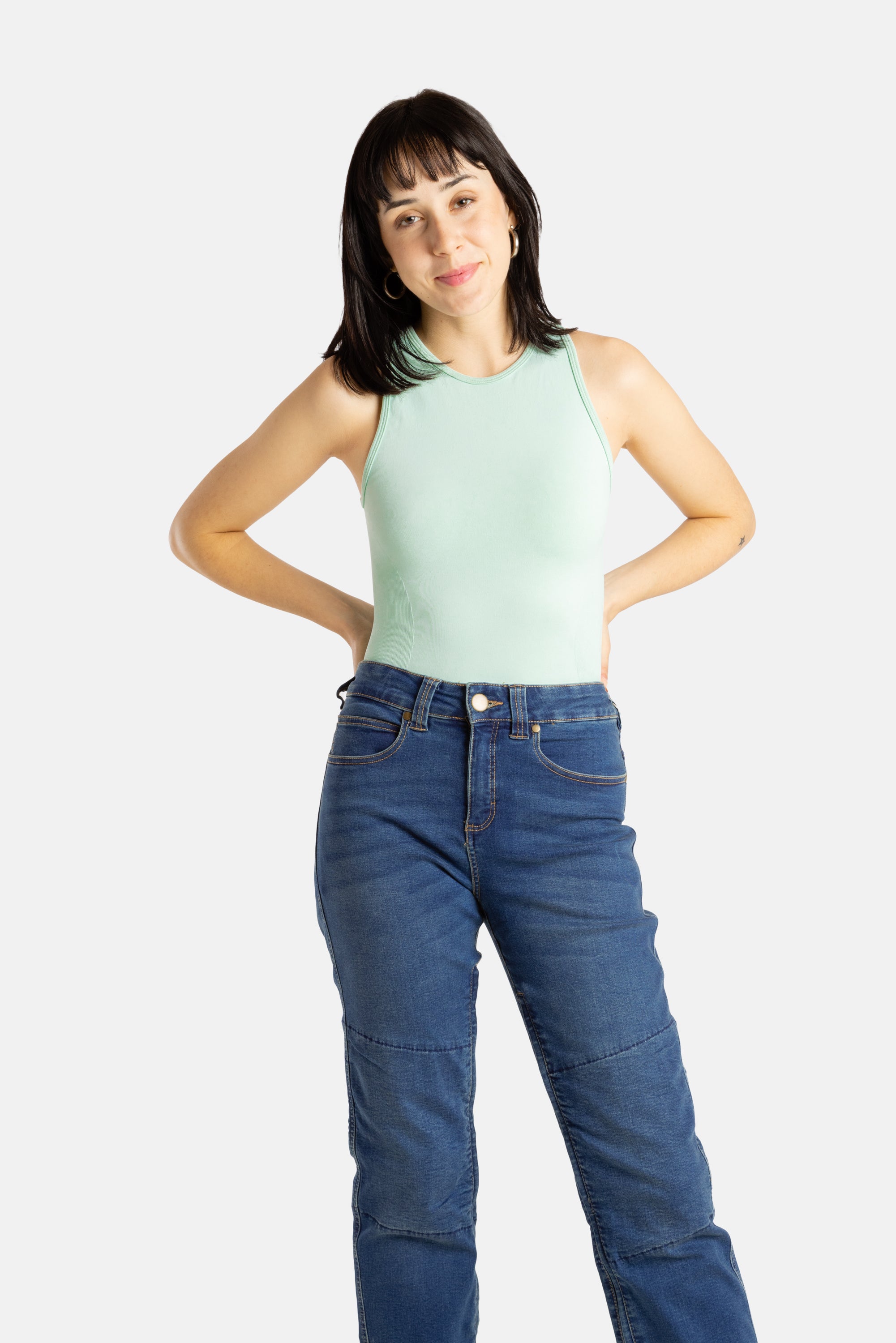 A white woman with long black hair and hoop earrings wears a sea form (Very light green) tank top and denim jeans.