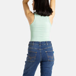 The back of a white woman with long black hair and hoop earrings wears a seaform (very light green) tank top and denim jeans.