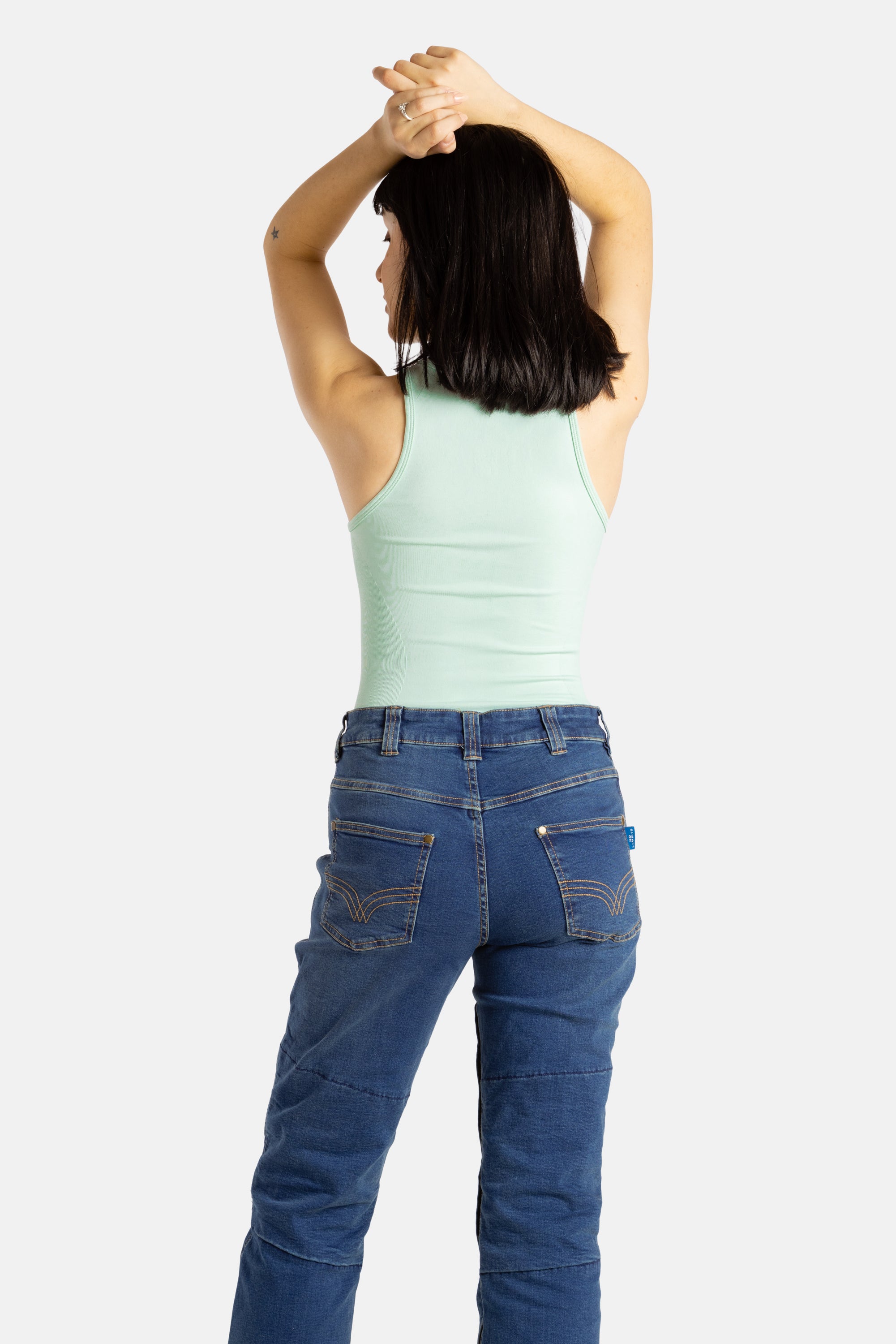 The back of a white woman with long black hair and hoop earrings wears a seaform (very light green) tank top and denim jeans.
