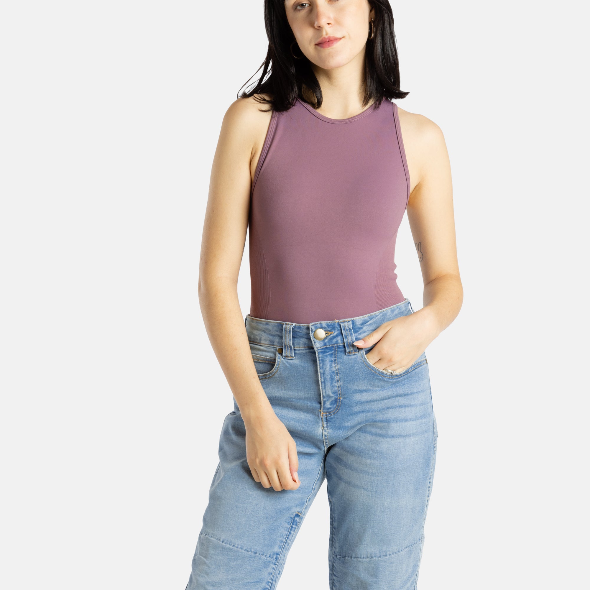A white woman with long black hair and hoop earrings wears a mauve tank top and denim jeans.
