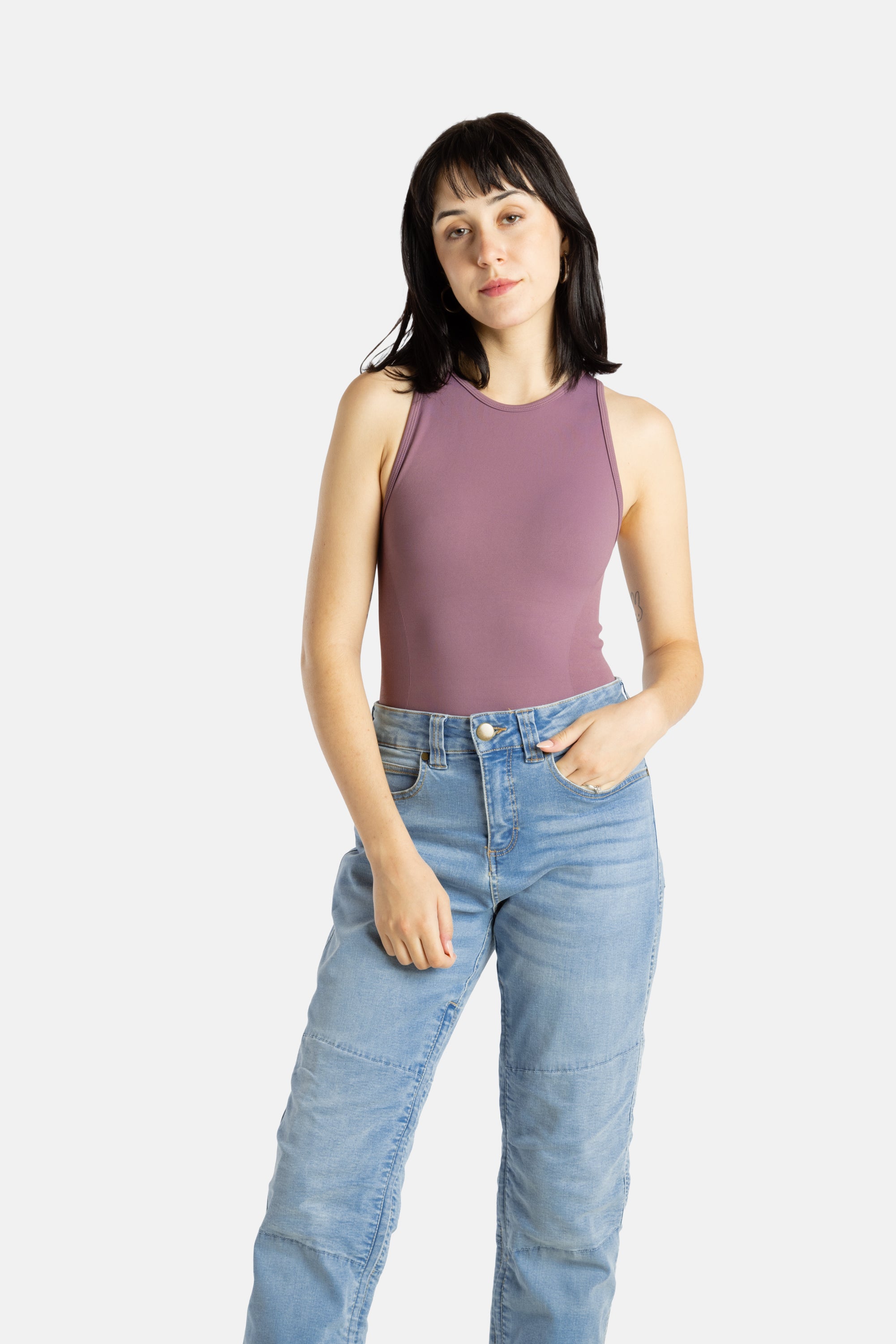 A white woman with long black hair and hoop earrings wears a mauve tank top and denim jeans.