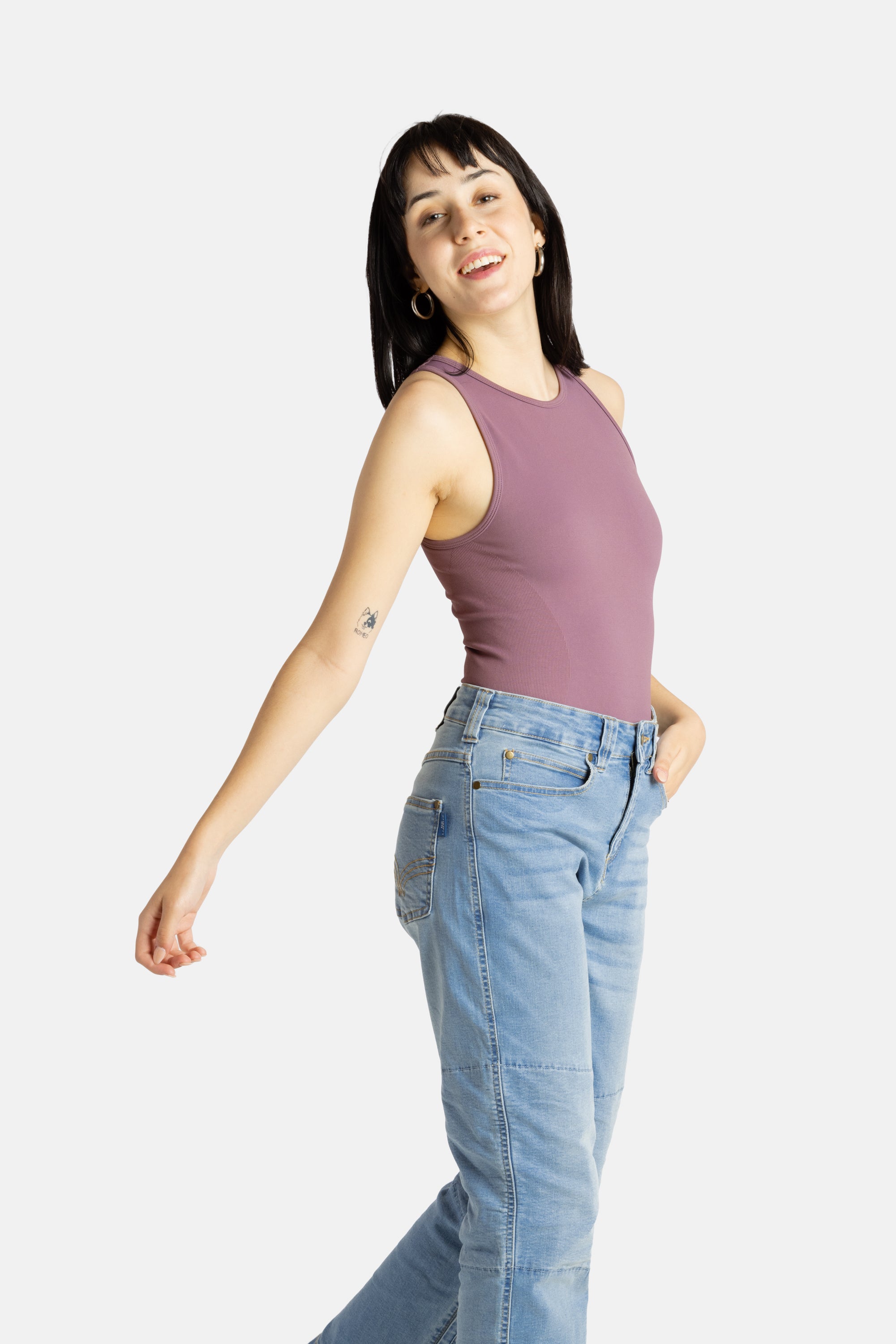 A white woman with long black hair and hoop earrings wears a mauve tank top and denim jeans.