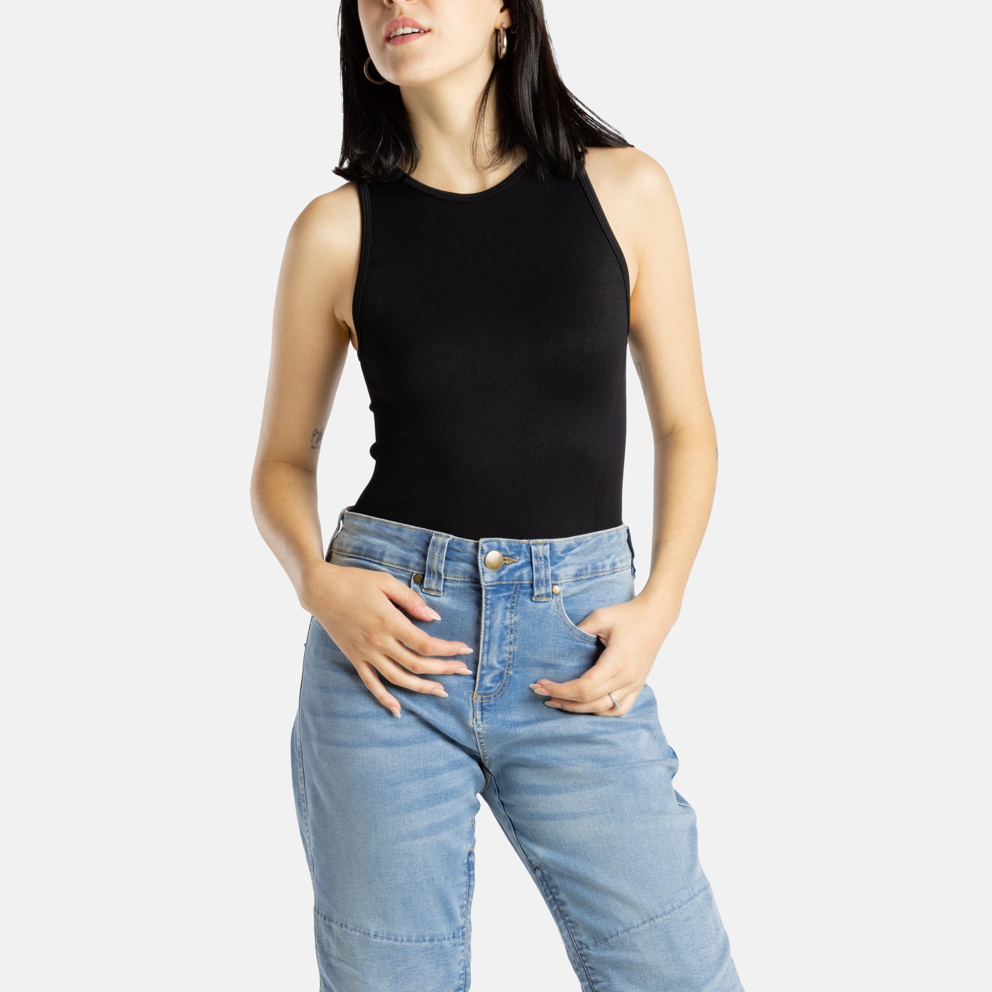 A white woman with long black hair and hoop earrings wears a black tank top and denim jeans.