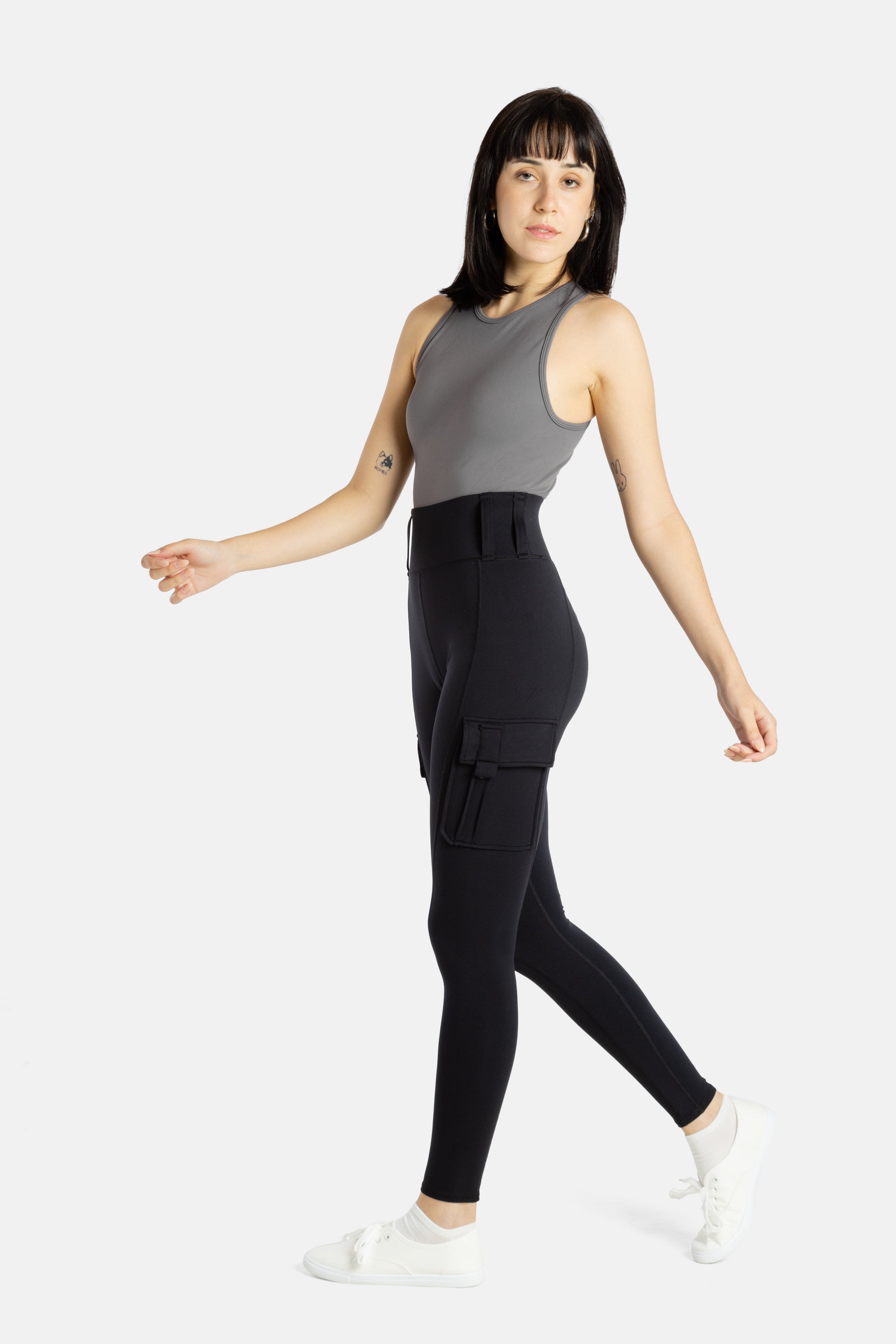 A white woman with long black hair and hoop earrings wears a charcoal tank top and black leggings.