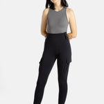 A white woman with long black hair and hoop earrings wears a charcoal tank top and black leggings. Her arms are behind her back and she raises her right ankle, showing her white sneakers.
