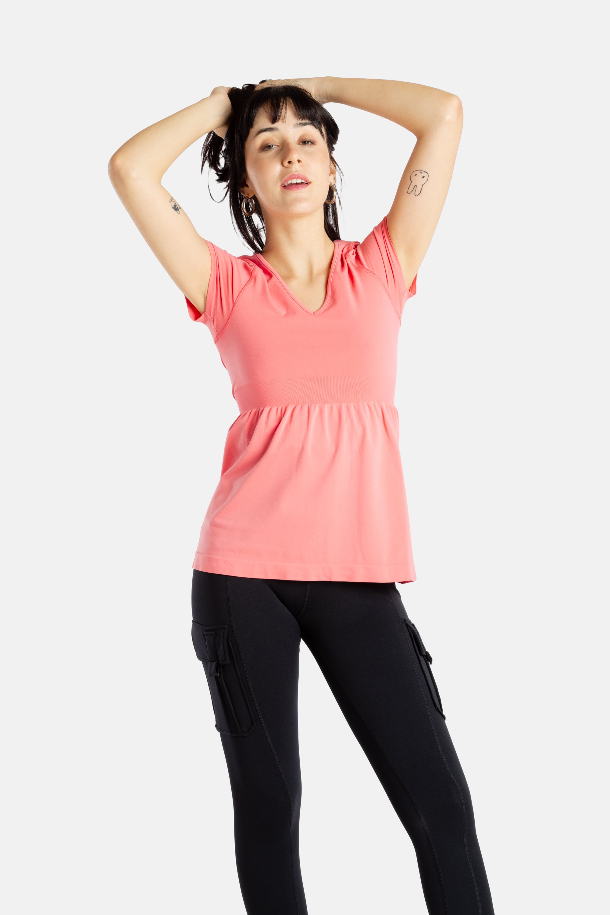 Sophia (A woman with long black hair) is wearing the No Limbits Adaptive Women's Coral Sensory Blouse with black leggings. She is pulling her hair back.