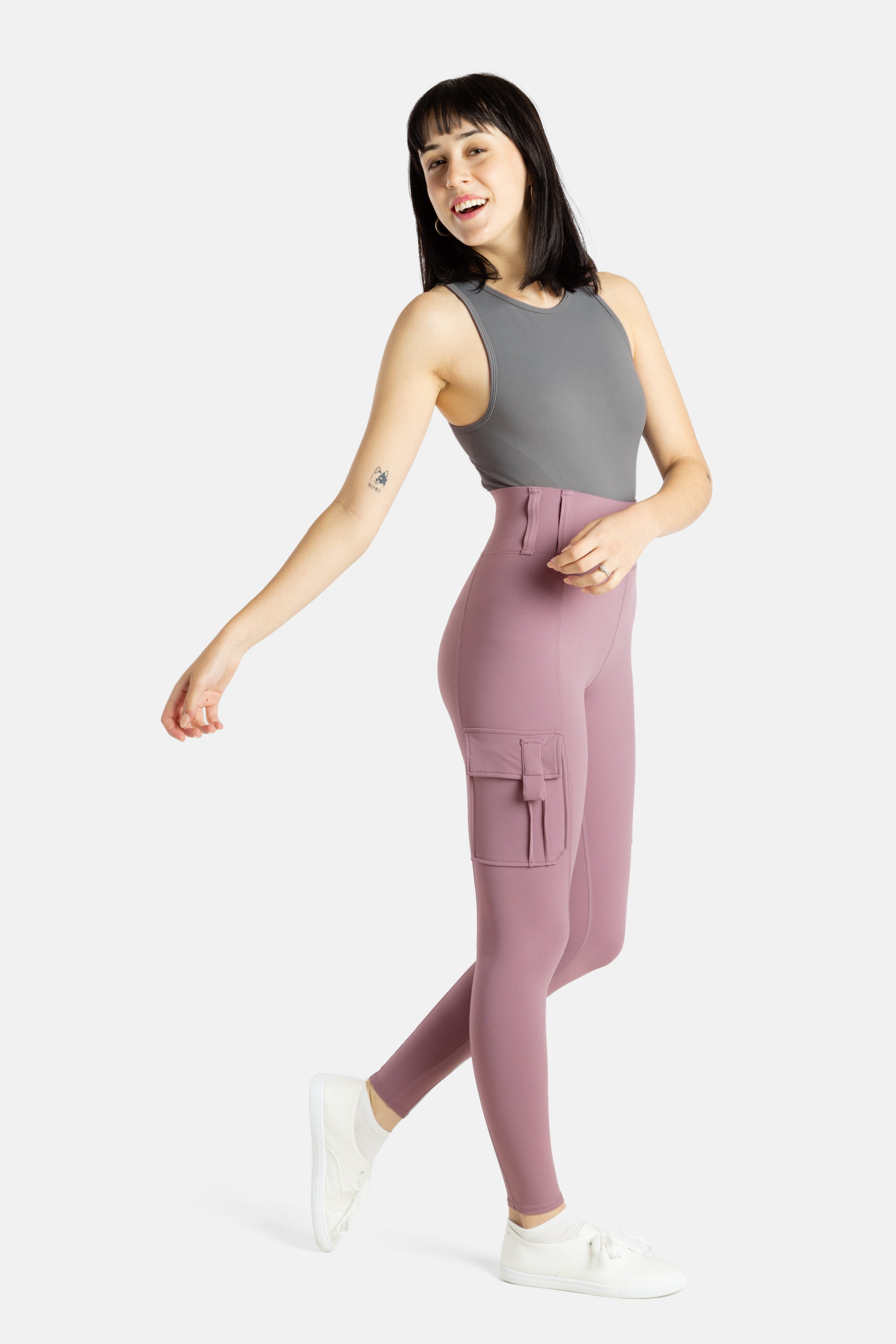 A white woman with long black hair and hoop earrings wears a charcoal tank top and mauve (Pinkish lavender) leggings.
