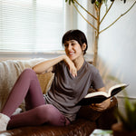 Sophia (A woman with long black hair) wears the No Limbits Adaptive Women's Charcoal Sensory Tee. She lays on a couch, holding a book.