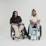 Princess, a Black woman on a pink wheelchair (with long locs, light pink lipstick, clear black sunglasses, and hoops) and Jessi, a Black man on a white wheelchair (with short locs with a bun on the top of his hair, a goatee, cross earrings, and white sneakers) hang out together and demonstrate the wheelchair pants.