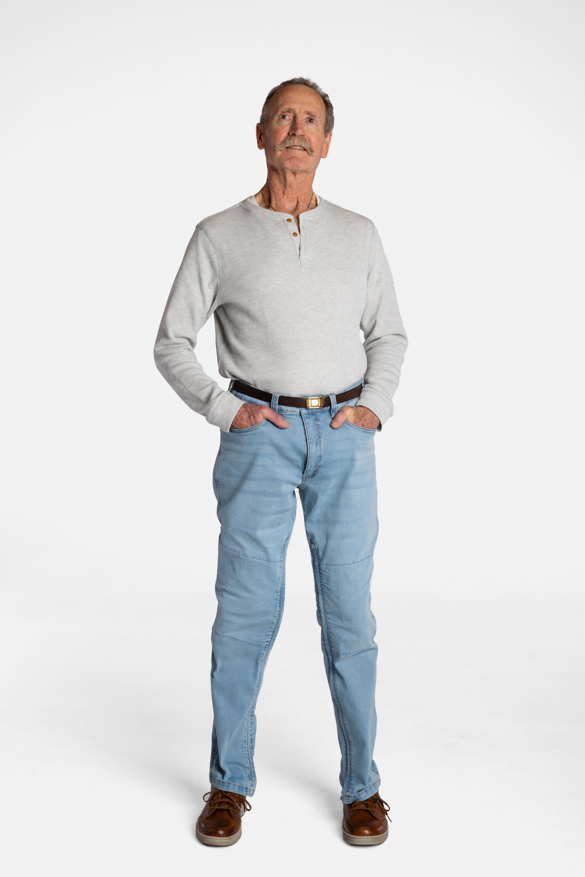 A white, elderly man with a mustache wears the Men’s Light Wash Unlimbited Pants, with a long-sleeved gray shirt, a brown belt, and brown shoes.