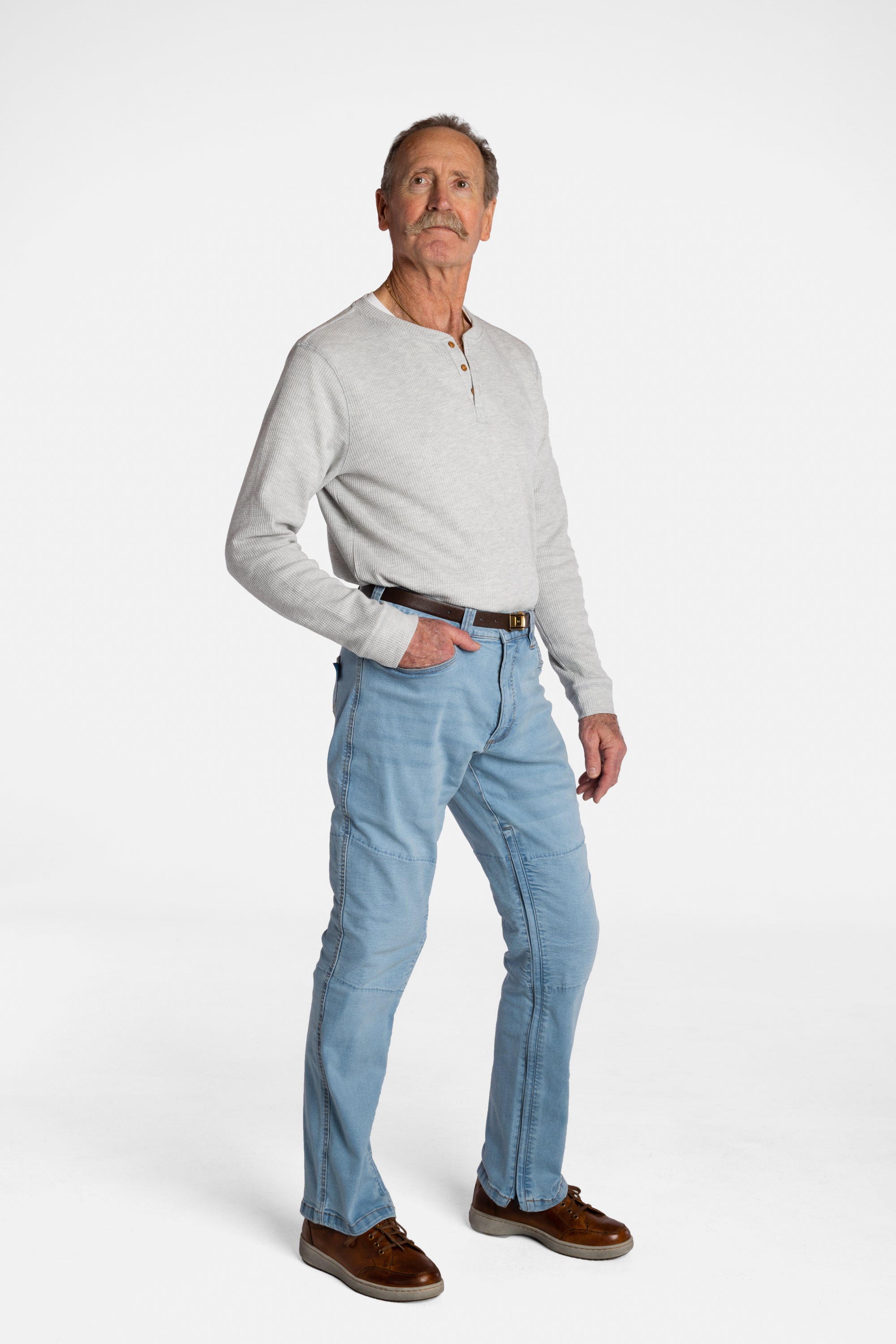 A white, elderly man with a mustache wears the Men’s Light Wash Unlimbited Pants, with a long-sleeved gray shirt, a brown belt, and brown shoes.
