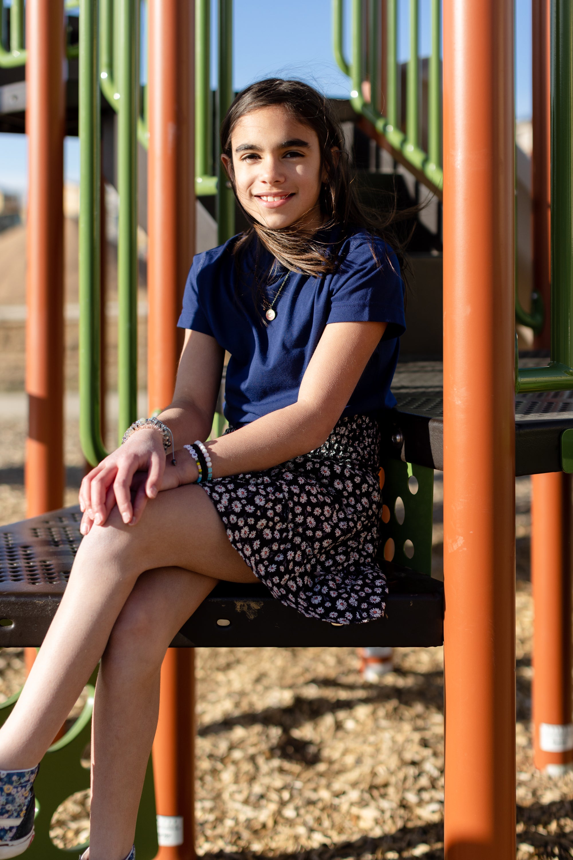A little girl is wearing a navy blue crewneck t-shirt. She is wearing a black and white skirt with black and white shoes. She is also sitting on a jungle gym. The colors of the jungle gym are orange, green and black.