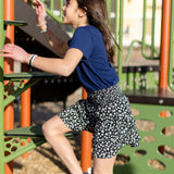 A little girl is wearing a navy blue crewneck t-shirt. She is wearing a black and white skirt with black and white shoes. She is also climbing on a jungle gym. The colors of the jungle gym are orange, green and black.
