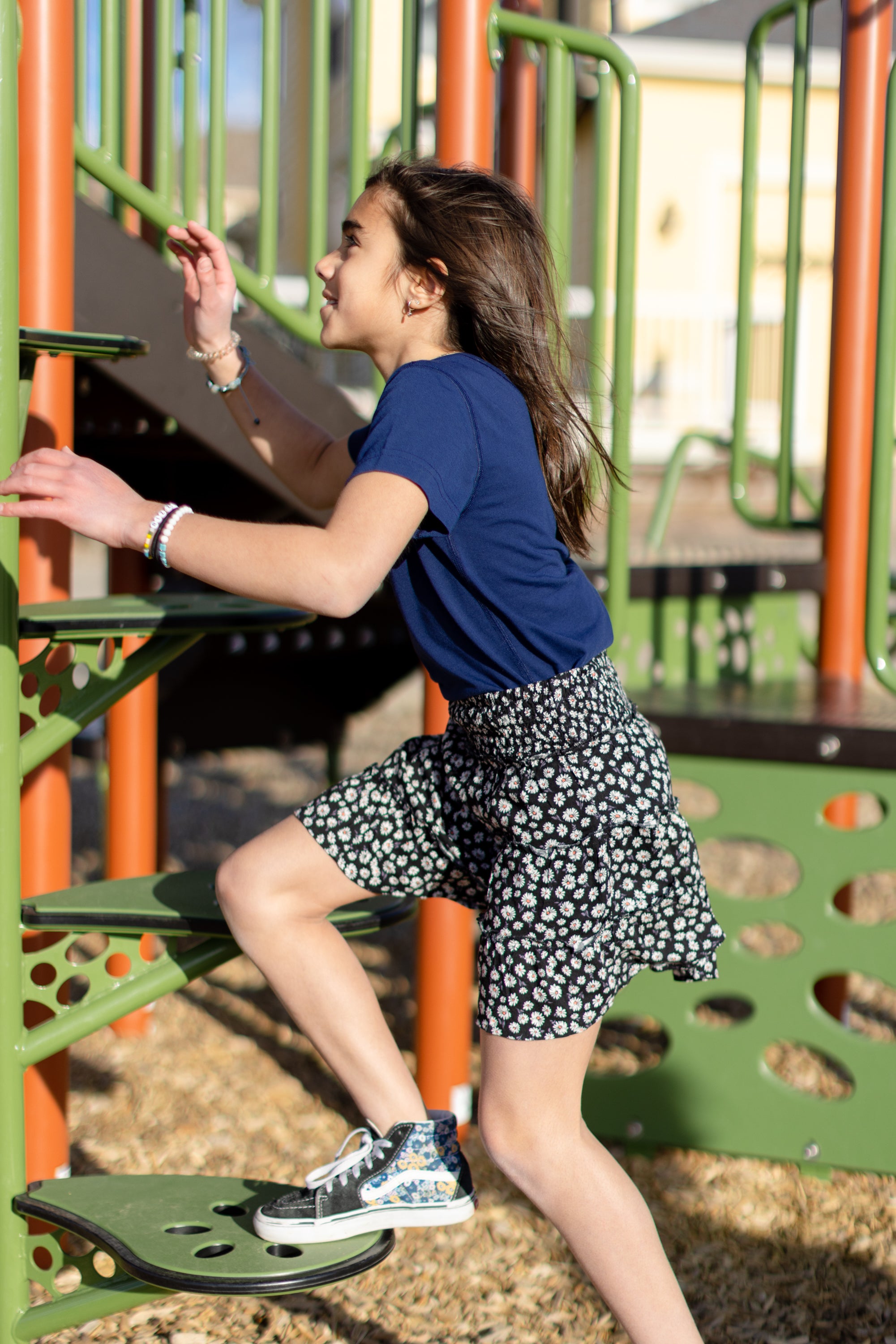 A little girl is wearing a navy blue crewneck t-shirt. She is wearing a black and white skirt with black and white shoes. She is also climbing on a jungle gym. The colors of the jungle gym are orange, green and black.