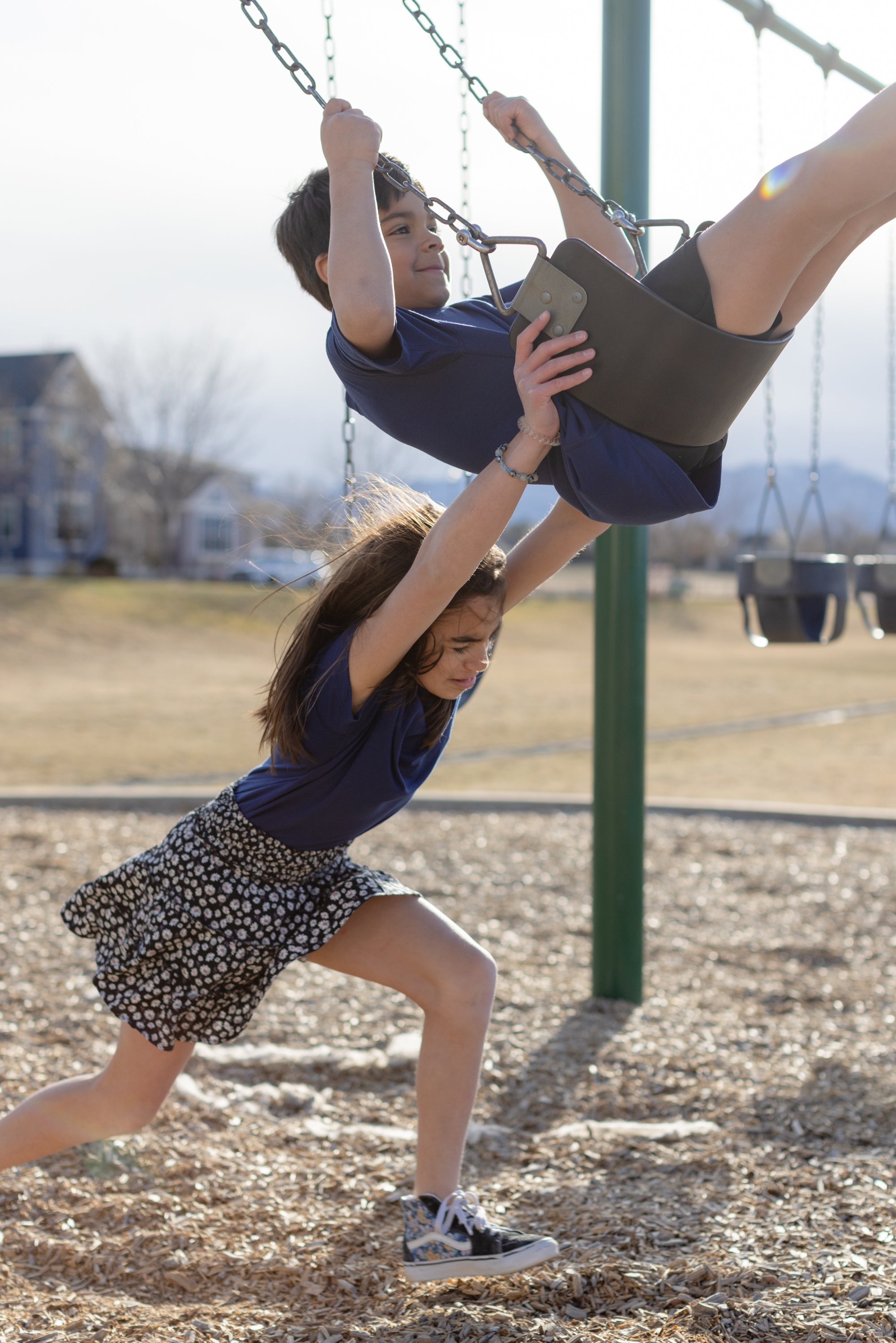 Two kids are playing on the swings together. The little girl is pushing the little boy who is sitting on the black swings. They are both wearing a navy blue basic t-shirt. The little girl is wearing a black and white skirt, and the boy is wearing black shorts.