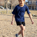 A little boy playing in the park, he is wearing a navy basic crewneck t-shirt and black shorts. You can see swing sets blurred out in the background of the photo.