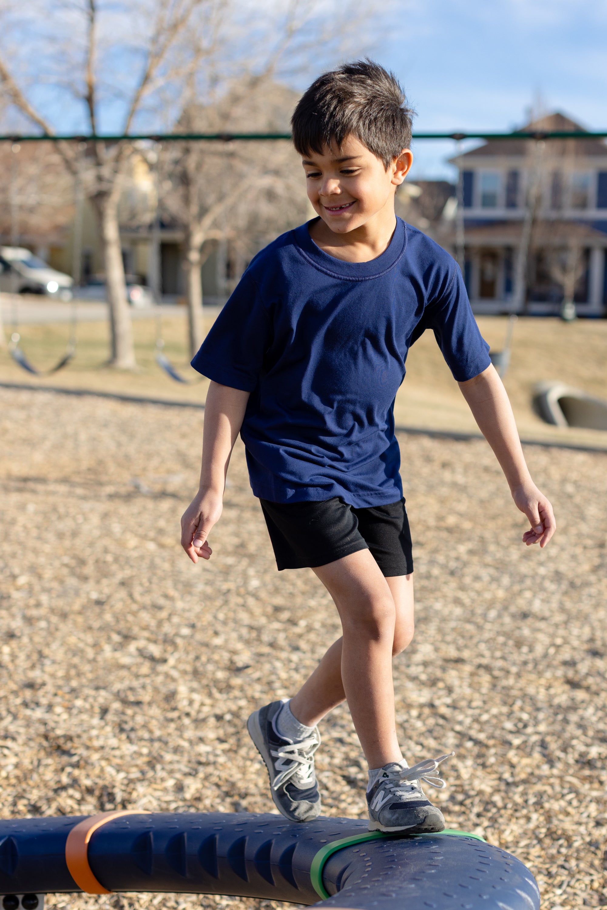A little boy playing in the park, he is wearing a navy basic crewneck t-shirt and black shorts. You can see swing sets blurred out in the background of the photo.