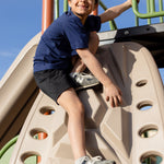 A little boy playing at the playground on a jungle gym climbing. He is wearing a navy blue basic t-shirt and black shorts. He has black hair and is looking off into the distance smiling.