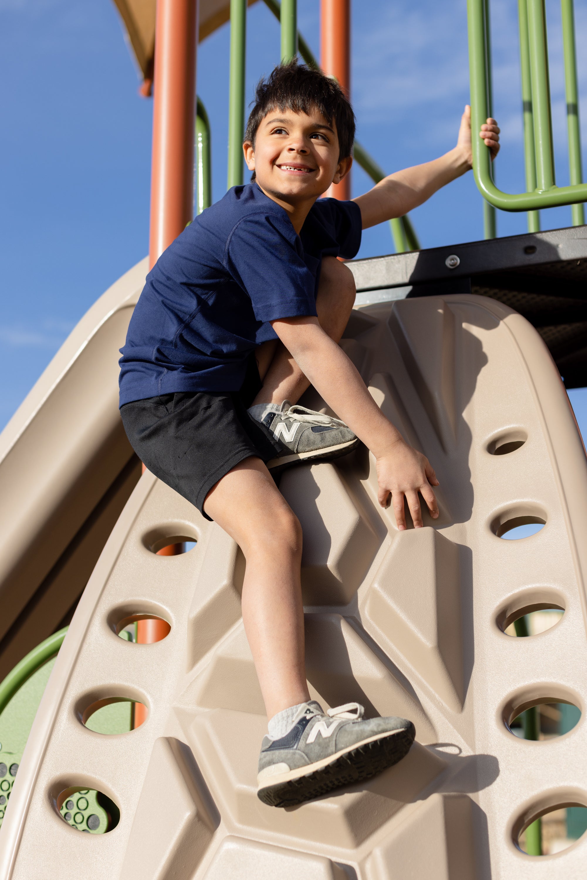 A little boy playing at the playground on a jungle gym climbing. He is wearing a navy blue basic t-shirt and black shorts. He has black hair and is looking off into the distance smiling.