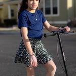 A little girl is wearing a basic navy t-shirt and a black and white skirt. She is standing on a purple and black skooter.