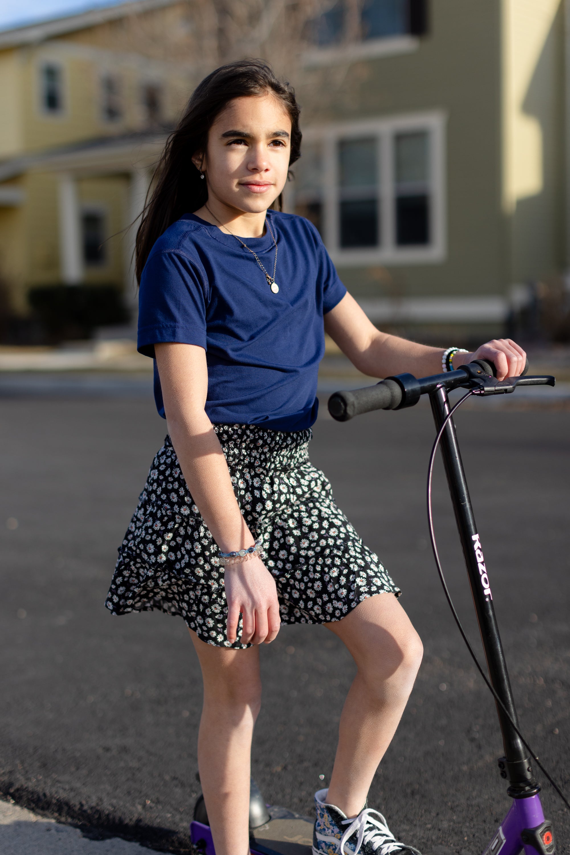 A little girl is wearing a basic navy t-shirt and a black and white skirt. She is standing on a purple and black skooter.