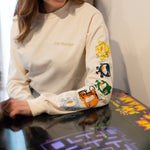 A blonde hair women wearing a cream long sleeve shirt. The cream long sleeve reads 'you matter' on the standard pocket area of the t-shirt. Running down the sleeve is multiple characters: A yellow one, navy, green, red, and light blue. The woman is sitting down playing a vintage game.