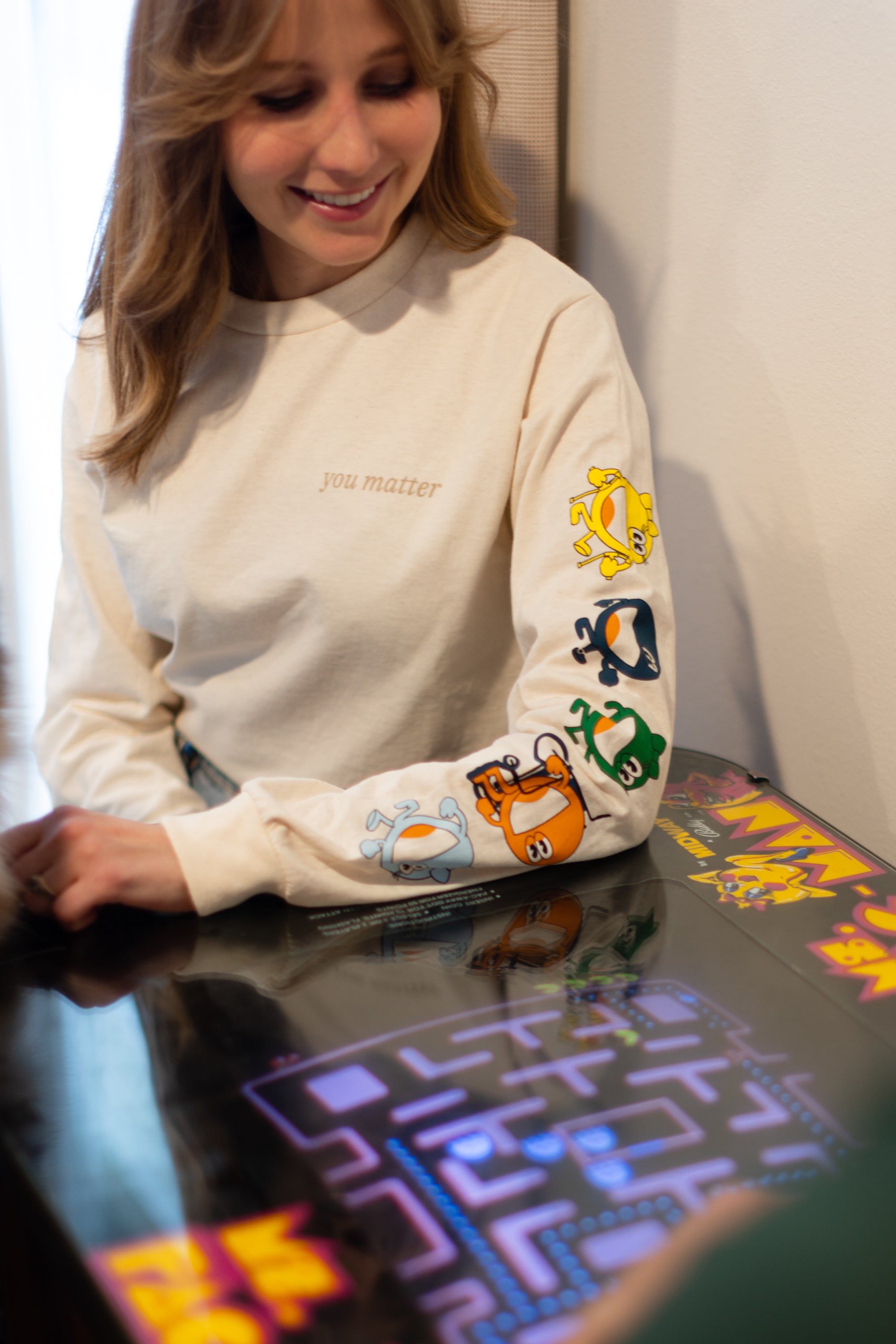 A blonde hair women wearing a cream long sleeve shirt. The cream long sleeve reads 'you matter' on the standard pocket area of the t-shirt. Running down the sleeve is multiple characters: A yellow one, navy, green, red, and light blue. The woman is sitting down playing a vintage game.