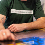 A white man (with a brown beard) wears a forest green short sleeve tee has a beige block printed on the front chest. The words "Not All Disabilities are Visible" are on this block. He is sitting and leans on a table.