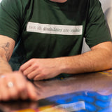 A white man (with a brown beard) wears a forest green short sleeve tee has a beige block printed on the front chest. The words "Not All Disabilities are Visible" are on this block. He is sitting and leans on a table.