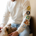 A man is sitting down by a window petting a dog. He is wearing a cream long sleeve shirt that reads 'you matter' on a t-shirt standard pocket area. Running down the sleeve are characters walking down, they are a circle shape. The colors of the characters are yellow, navy, green, orange, and light blue.