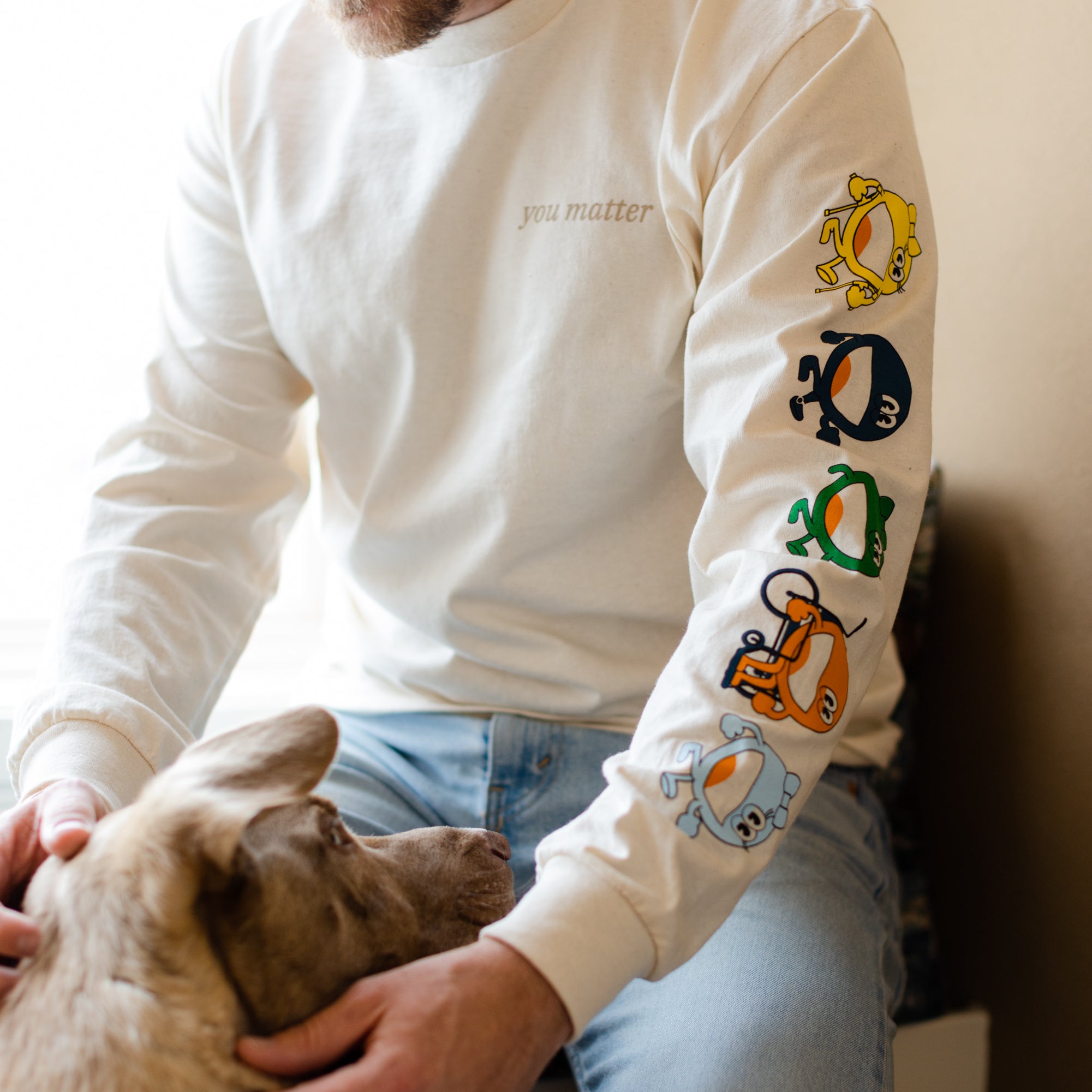 A man is sitting down by a window petting a dog. He is wearing a cream long sleeve shirt that reads 'you matter' on a t-shirt standard pocket area. Running down the sleeve are characters walking down, they are a circle shape. The colors of the characters are yellow, navy, green, orange, and light blue.