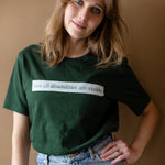 A white woman (with long brown hair) wears a forest green short sleeve tee has a beige block printed on the front chest. The words "Not All Disabilities are Visible" are on this block.