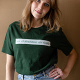 A white woman (with long brown hair) wears a forest green short sleeve tee has a beige block printed on the front chest. The words "Not All Disabilities are Visible" are on this block.