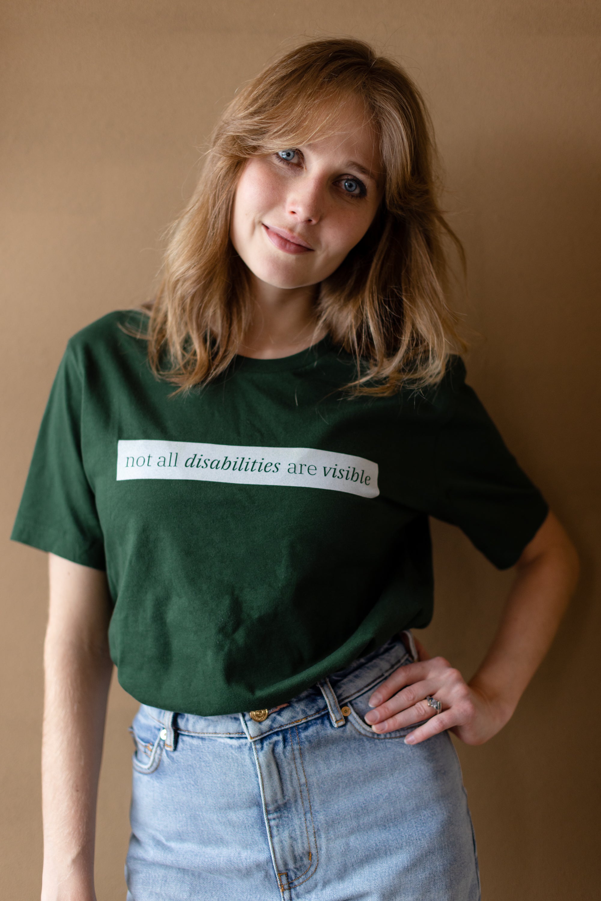 A white woman (with long brown hair) wears a forest green short sleeve tee has a beige block printed on the front chest. The words "Not All Disabilities are Visible" are on this block.