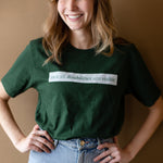 A white woman (with long brown hair) wears a forest green short sleeve tee has a beige block printed on the front chest. The words "Not All Disabilities are Visible" are on this block.