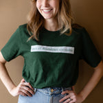 A white woman (with long brown hair) wears a forest green short sleeve tee has a beige block printed on the front chest. The words "Not All Disabilities are Visible" are on this block. 