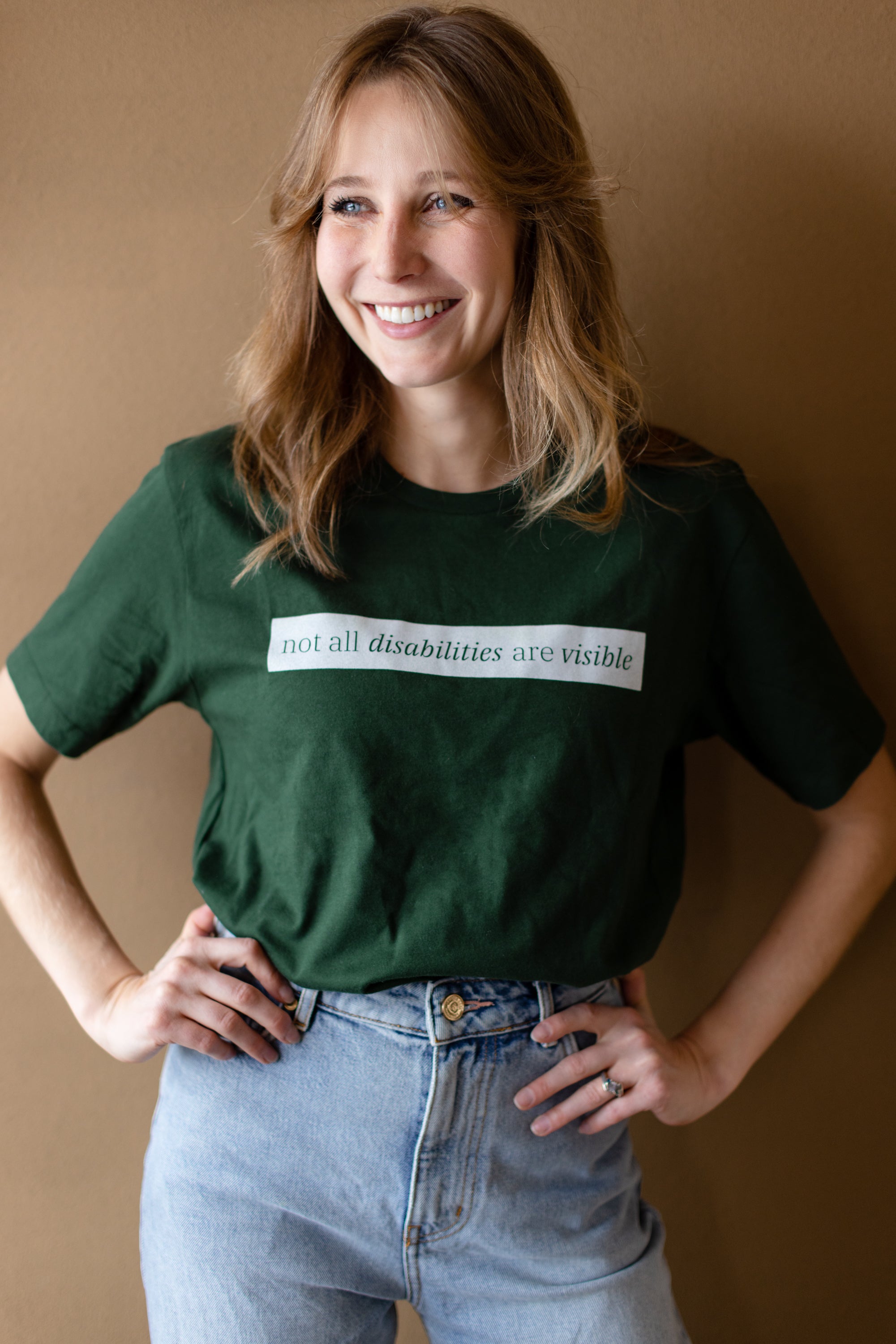 A white woman (with long brown hair) wears a forest green short sleeve tee has a beige block printed on the front chest. The words "Not All Disabilities are Visible" are on this block.