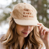 A white woman with long brown hair wears a cream baseball cap with brown text that says "disability is not a bad word."