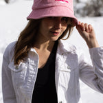 A white woman with brown long hair wears a light pink bucket hat with a white illustration of the sun and pink text that says "Unlimbited." She's in a snowy forest.