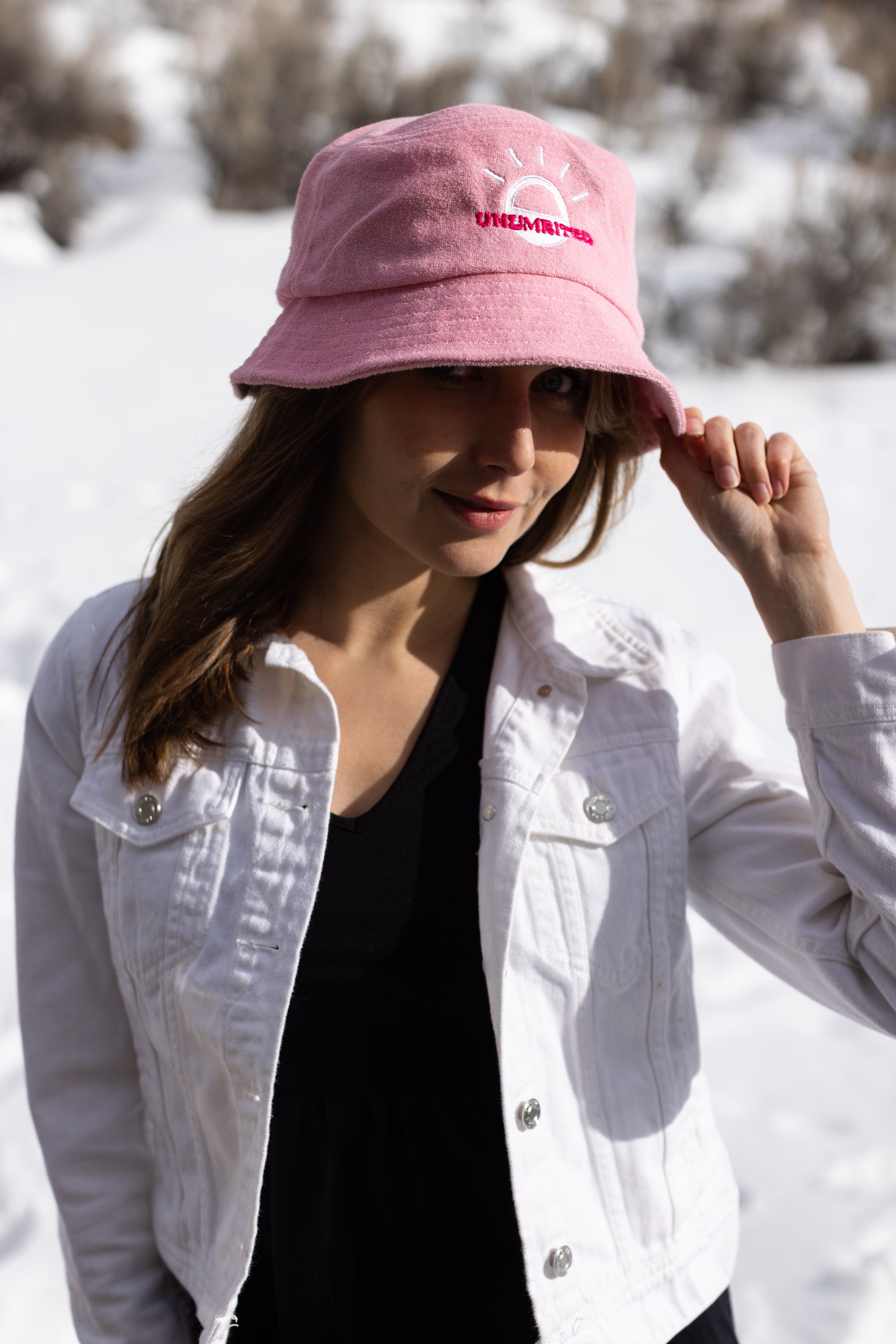 A white woman with brown long hair wears a light pink bucket hat with a white illustration of the sun and pink text that says "Unlimbited." She's in a snowy forest.