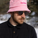 A white man with a beard and brown sunglasses wears a light pink bucket hat with a white illustration of the sun and pink text that says "Unlimbited." He's in a snowy forest.
